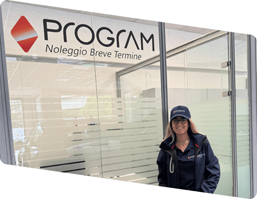 Donna con cappellino e giacca aziendale davanti all’ufficio noleggio Program Noleggio Breve Termine Valdarno, con insegna e vetrata visibili.